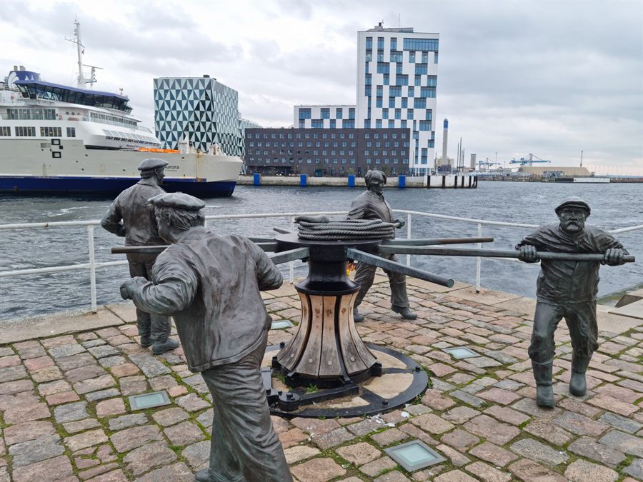 Bronze sculpture of sailors heaving on a capstan on the Copenhagen waterfront, a ferry behind